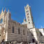 A grand, intricately detailed cathedral, possibly the Siena Duomo, stands against a clear, vibrant blue sky on a sunny day. The lower portion shows a crowd of people gathered around the base of the building, with some colorful flags visible.
