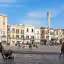 An editorial style image showcasing a bustling city square in Italy, featuring historic buildings, a prominent column, and people walking about under a clear blue sky.