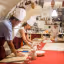 A group of people wearing chef hats and aprons are engaged in a cooking class, rolling dough on a large red table in a rustic kitchen setting.