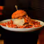 A professionally taken close-up image of a large burger with sweet potato fries, presented on a white plate and held by a person in a dark uniform. The background is dark and out of focus, highlighting the food.