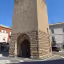 A well-lit and sharp photograph of an old stone tower with an archway, set against a clear blue sky. The image captures the historical details of the structure and appears highly photo-realistic.