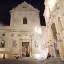 An evening view of a grand, ornately designed Baroque church facade, illuminated by artificial lights against a dark night sky, with a few people gathered in the plaza below.