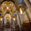 An interior shot of an ornate church or cathedral, featuring intricate gold mosaics covering the vaulted ceilings and walls. Large marble pillars frame the central view towards the altar. The lighting appears natural, highlighting the rich details of the space.