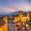An elevated view of a lively town square and surrounding buildings at twilight. The scene features a prominent clock tower, a church roof in the foreground, and a bustling square filled with people. In the background, a coastline extends towards mountains under a colorful sky.