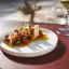 A professionally taken close-up image of a gourmet seafood dish, likely fried octopus or similar small seafood, artfully arranged with crispy elements and vibrant red and green sauces on a white plate. The background is blurred, showing an outdoor or window setting.