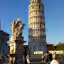 The image prominently features the Leaning Tower of Pisa and a classical statue against a clear blue sky. The composition effectively highlights the iconic landmark and the foreground sculpture, both rendered with excellent sharpness and detail. The natural lighting is superb, illuminating the stone structures and providing accurate, vibrant colors. The image feels authentic and photo-realistic, with all key elements in clear focus.