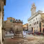 An image of a historic town square featuring a central well, traditional stone buildings, and a clear blue sky. The composition is balanced, capturing various elements of the square including people and outdoor seating areas.