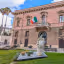 A well-composed and brightly lit photograph showcasing a pink historical building with an Italian flag, a prominent palm tree, and a decorative statue in the foreground. The image displays high clarity and natural colors.
