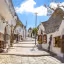 An editorial-style image of a charming, narrow street lined with traditional white-washed houses featuring unique cone-shaped stone roofs, characteristic of Alberobello, Italy. The street is paved with stones and slopes upwards, leading the eye through the scene. The sky is bright blue with some clouds.