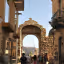 An editorial-style image capturing a street scene in a historic town, featuring an ancient stone archway framing a view of distant mountains and a church dome, with people walking through the street.