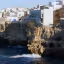 The image is a well-composed and vibrant depiction of a coastal town situated atop rugged cliffs overlooking the sea. The natural lighting is excellent, illuminating the scene effectively and enhancing the textures of the buildings and cliffs. The image is sharp with clear details, and the colors, particularly the blues of the sea and the whites of the buildings, appear accurate and natural. The photograph is highly realistic, and all key elements are in sharp focus.