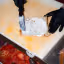 An overhead shot of hands in black gloves spreading a white creamy substance, likely cream cheese, onto a piece of bread or base on a light-colored cutting board. Other ingredients like sliced tomatoes are visible in the background.
