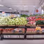 The image is well-composed, providing a clear and comprehensive view of a supermarket's produce section. It benefits from excellent lighting, which highlights the vibrant and natural colors of the fresh produce, contributing to its strong photo-realistic quality. Key elements like the fruits and vegetables in the foreground and mid-ground are sharp and in focus. While standard price tags and background store signage are present, they are integral to an editorial depiction of a retail environment and do not detract from the overall quality or function as primary promotional graphics.