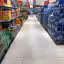 An interior shot of a supermarket aisle, featuring shelves stocked with various bottled beverages and water. The aisle floor is clean and clear, with the view extending towards the back of the store where a shopping cart and a person are faintly visible.