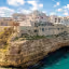 A beautiful panoramic view of a coastal town built atop rugged cliffs, overlooking a stunning turquoise sea under a bright blue sky with scattered white clouds. The ancient buildings with their white and pastel facades contrast beautifully with the vibrant water.