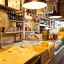 An inviting interior shot of a rustic shop or bar, featuring shelves laden with bottles, glassware, and an antique Berkel scale in the foreground. The warm lighting highlights the various products and the rich texture of the wooden shelves and counter.