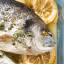 A professionally taken close-up of a cooked whole fish, garnished with green herbs and lemon slices, served in a clear glass baking dish.