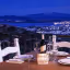 An evening shot of a table set for dinner on a balcony or terrace, overlooking a city with twinkling lights, a body of water, and mountains in the background, all bathed in the blue hour light.