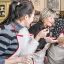 An image capturing three women engaged in cooking within a kitchen setting, dressed in aprons. One woman is stirring a pan on the stove, while the others observe and interact. The image conveys a sense of communal activity and learning.