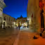 A night-time view of an old town street, featuring historic buildings illuminated by artificial lights under a deep blue twilight sky. The street, paved with light-colored stones, leads towards an archway in the distance. People are present on the street, with a street musician visible on the right.