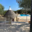 The image is sharp and photo-realistic, accurately capturing the colors of the outdoor scene under bright sunlight. The key elements, including the Trullo and the patio furniture, are clearly in focus. The composition effectively frames the Trullo and integrates the pool area into the shot.