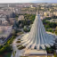 An excellent aerial shot showcasing the unique, modern architecture of the Sanctuary of Our Lady of Tears in Syracuse. The composition is strong, centering the distinctive conical structure against a backdrop of the city.