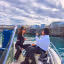 An image capturing two women on a boat enjoying the view of a coastal town, with one person taking a photo of the other. The scene is bright and shows clear blue water and a cloudy sky.