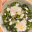 A close-up, top-down view of a savory breakfast dish featuring two eggs cooked in a bed of green spinach and peas, topped with crumbled white feta cheese. The dish is presented in a light-colored bowl.