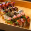 An editorial-style image showcasing a variety of intricately prepared sushi rolls presented in a rustic wooden tray. The rolls feature diverse toppings and garnishes, including various types of fish, avocado, herbs, and edible flowers, arranged artfully to highlight their details and textures.