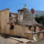 An exterior view of a traditional Trullo house with its distinctive conical stone roof, set on a sunny street with a stone wall adorned with red flowers.