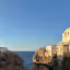 An outdoor, eye-level, wide shot of a scenic Italian coastal town built into cliffs overlooking a popular beach cove during what appears to be golden hour. The clear blue sky meets the horizon of the calm sea. People are visible on the sandy beach below the rugged cliffs.