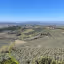 A panoramic view of a vast, sunny countryside featuring rolling hills, extensive olive groves, vineyards, and distant mountains under a clear blue sky.