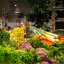 An editorial style image showcasing a vibrant and diverse selection of fresh vegetables, including various types of lettuce, purple and green cauliflower, carrots, and leeks, arranged appealingly on a display. The foreground is in sharp focus, while the background, depicting a blurred retail environment with ambient lighting, provides context.