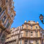 An eye-level shot of ornate historical architecture against a clear blue sky, featuring a prominent, detailed building with statues and balconies, flanked by another building and a decorative street lamp.
