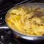 A close-up shot of broad, flat pasta, likely pappardelle, in a pan with a meat-based sauce, simmering on a stove with a visible flame.