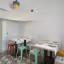 An interior shot of an empty restaurant dining area, featuring multiple tables set with place settings and colorful stools. The room has a white ceiling and walls, with a distinct patterned floor.