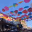 An outdoor market scene where numerous colorful umbrellas are suspended overhead, forming a vibrant canopy against a clear blue sky. Below, market stalls with various goods are visible, giving a sense of a lively public space.