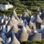 An elevated view showcasing a dense cluster of traditional trulli houses with their distinctive conical stone roofs, set amidst green foliage in Puglia, Italy.