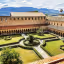 An editorial style image showcasing a beautiful and well-maintained historic courtyard with intricate garden design, surrounded by ancient monastic architecture, with mountains visible in the background under a clear sky.