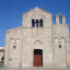 A well-lit, frontal view of an ancient stone church under a clear blue sky, showcasing its rustic stone facade, arched windows, and small bell tower.